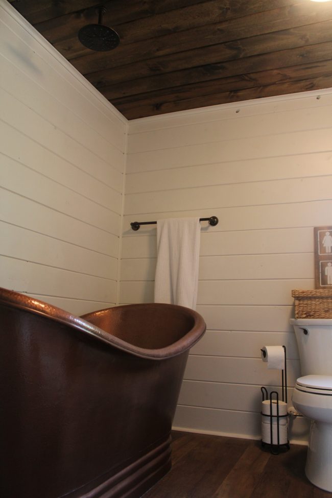 A modern farmhouse bathroom makeover - with a copper tub, copper sink, wood plank ceiling, white shiplap walls, LVT flooring, and granite counter tops.